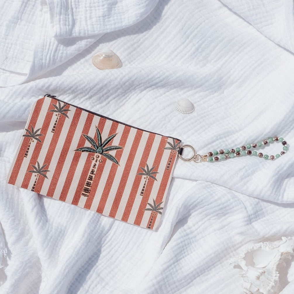 A rectangular clutch or wallet with red and white vertical stripes and palm tree motifs, resting on a crumpled white gauze fabric over a sandy brown ground.