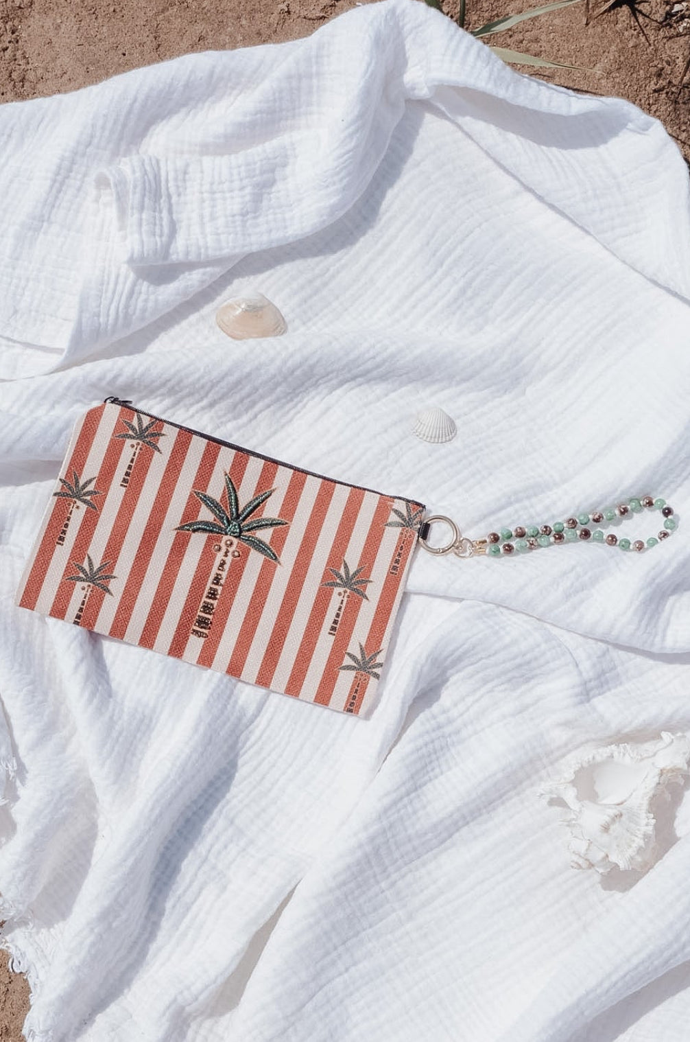 A rectangular clutch or wallet with red and white vertical stripes and palm tree motifs, resting on a crumpled white gauze fabric over a sandy brown ground.