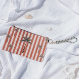 A rectangular clutch or wallet with red and white vertical stripes and palm tree motifs, resting on a crumpled white gauze fabric over a sandy brown ground.