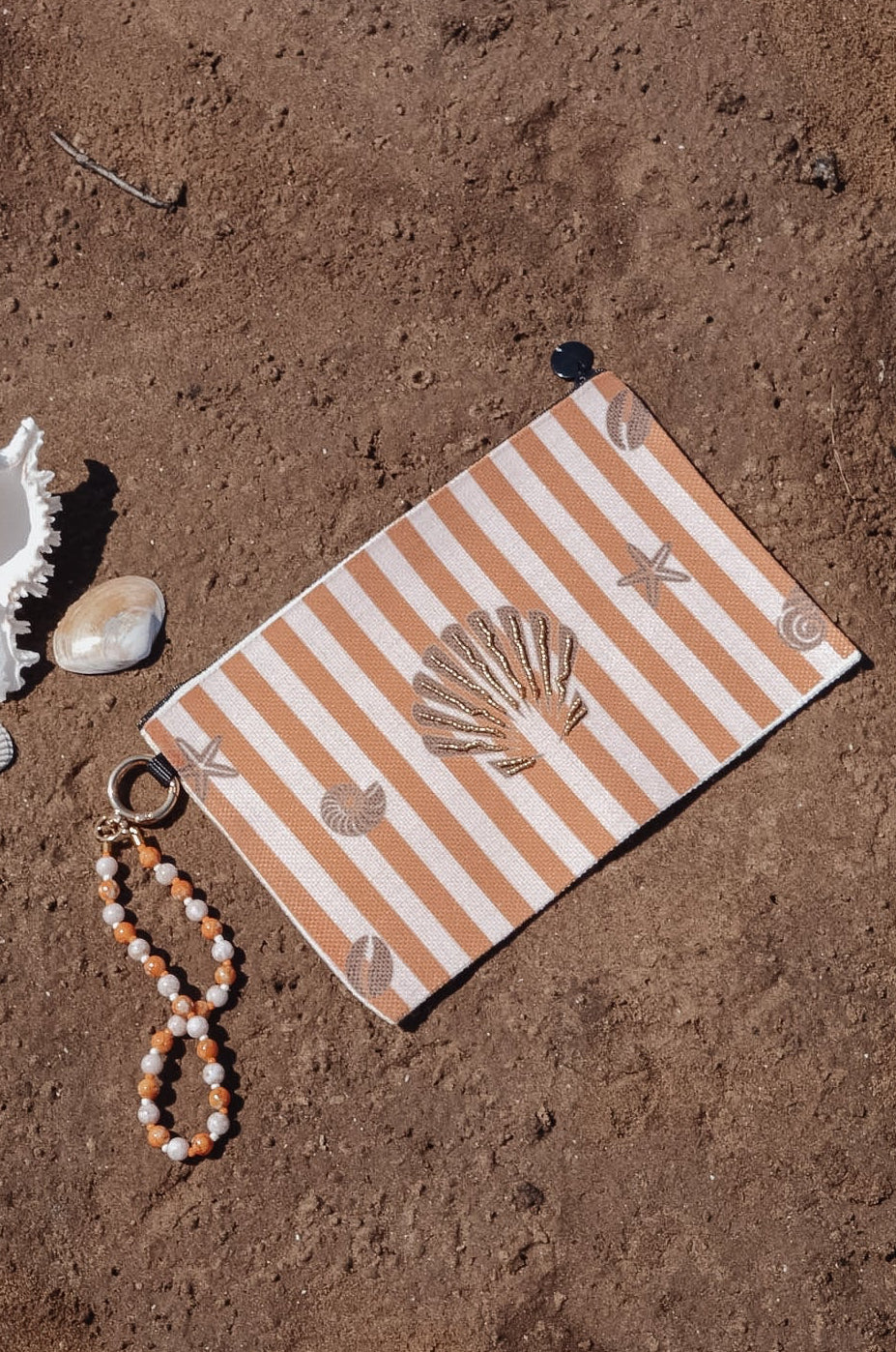 A beach-themed wristlet pouch with orange and white stripes and seashell motifs, styled on sandy ground with a white conch shell, a small bead strand, and a gold ring.