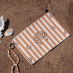 A beach-themed wristlet pouch with orange and white stripes and seashell motifs, styled on sandy ground with a white conch shell, a small bead strand, and a gold ring.