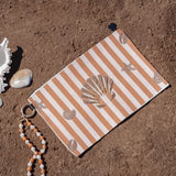 A beach-themed wristlet pouch with orange and white stripes and seashell motifs, styled on sandy ground with a white conch shell, a small bead strand, and a gold ring.