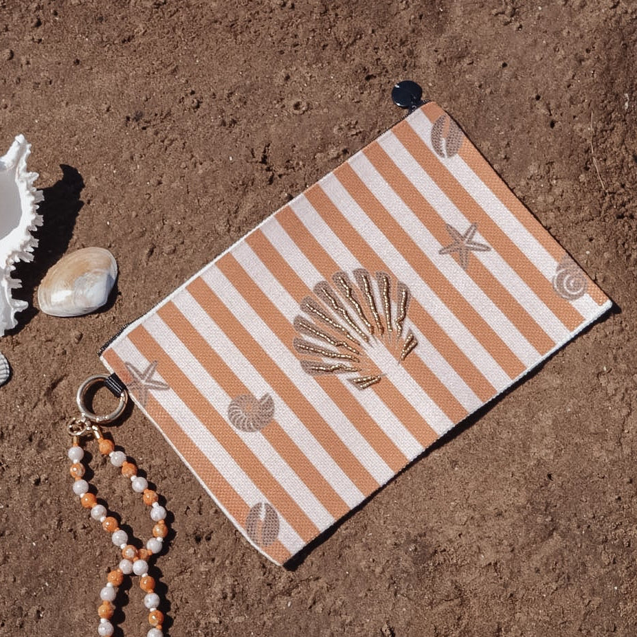 A beach-themed wristlet pouch with orange and white stripes and seashell motifs, styled on sandy ground with a white conch shell, a small bead strand, and a gold ring.